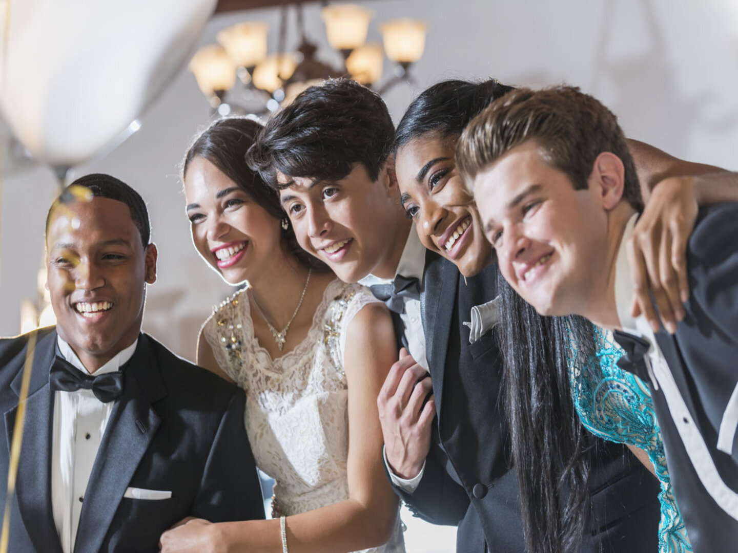 A group of five multi-ethnic teenagers and  young adults dressed in formalwear - dresses and tuxedos. They are at a special event, a prom or party at a restaurant.